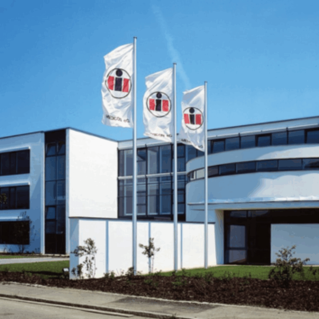 Medicon Gebäude im Gänsäcker 15 in Tuttlikngen. Modernes Büro- oder Firmengebäude mit großen Glasfenstern und weißer Fassade. Vor dem Gebäude wehen drei Fahnen mit einem rot-schwarz-weißen Firmenlogo auf hohen Fahnenmasten bei blauem Himmel.