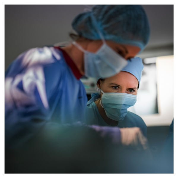 Excellence in Surgical Assistance_Medicon Courses. A close-up of a surgeon wearing surgical scrubs, including a surgical cap and a medical face mask, with a focused expression. She is in an operating room and is about to perform or assist with a procedure. In the foreground, another person in surgical scrubs is partially visible.