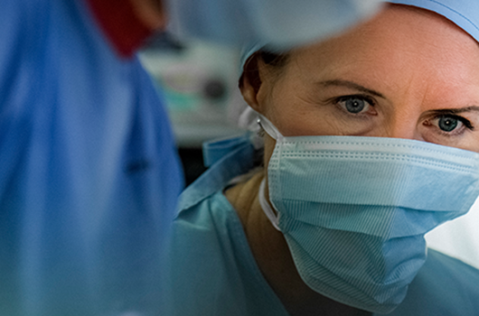 Titelbild für den Medicon Kurs Op-Excellence. Nahaufnahme einer konzentriert blickenden Chirurgin in OP-Kleidung, inklusive Haube und medizinischem Mund-Nasen-Schutz. Sie befindet sich in einem Operationssaal und ist im Begriff, einen Eingriff durchzuführen oder daran teilzunehmen. Im Vordergrund ist eine weitere Person in OP-Kleidung teilweise sichtbar.